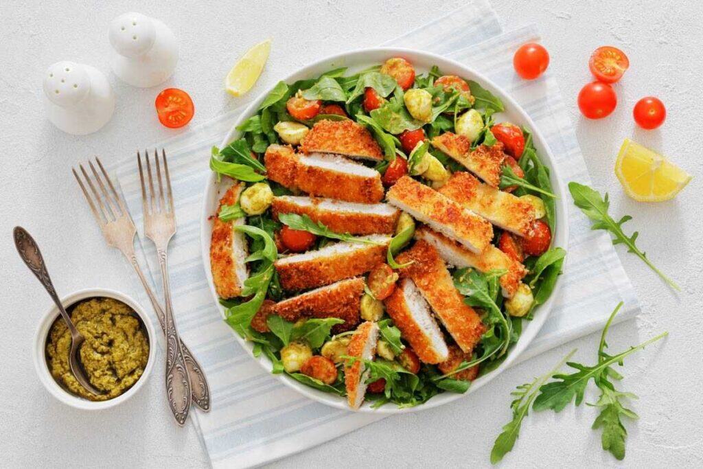 Fully plated Chicken Parmesan Salad on a white plate, with crispy chicken, cherry tomatoes, arugula, and bocconcini, served alongside a small bowl of pest