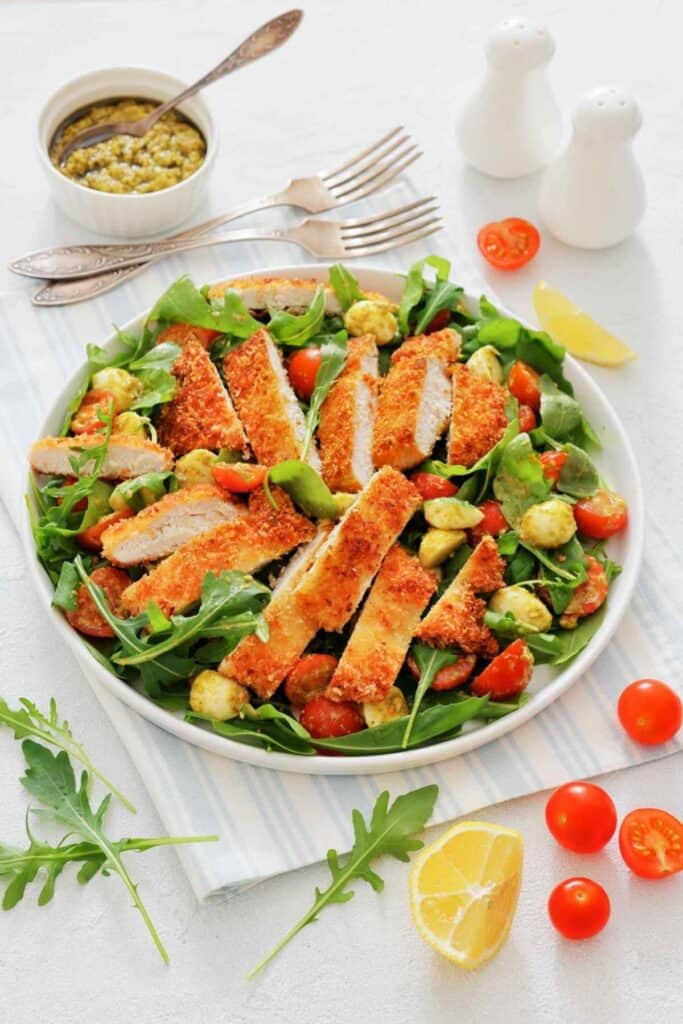 A white plate is filled with a vibrant Chicken Parmesan Salad. Crispy, golden chicken cutlets rest on top of fresh arugula, with cherry tomatoes and creamy bocconcini scattered around.