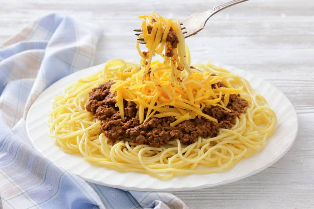 A plate of spaghetti covered with Cincinnati chili, topped with shredded cheddar cheese and chopped onions, with a fork lifting a bite.