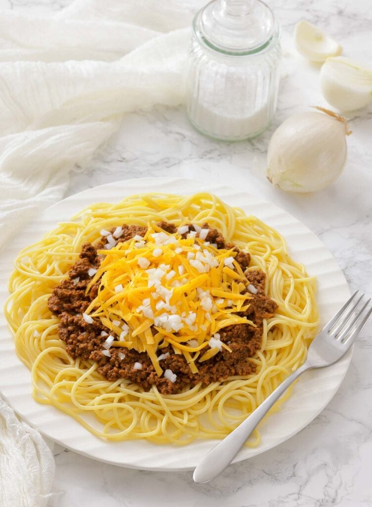 A plate of spaghetti topped with meat sauce and shredded cheddar cheese sits on a table next to a cloth napkin and a block of cheddar with a grater.