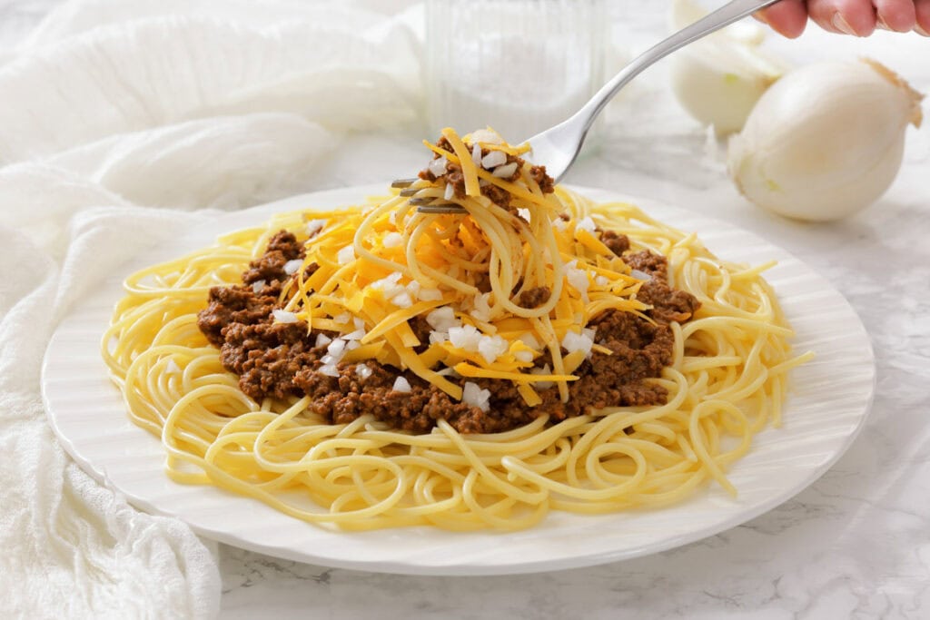 A plate of spaghetti topped with Cincinnati chili, shredded cheddar cheese, and chopped onions, with a fork lifting a bite.