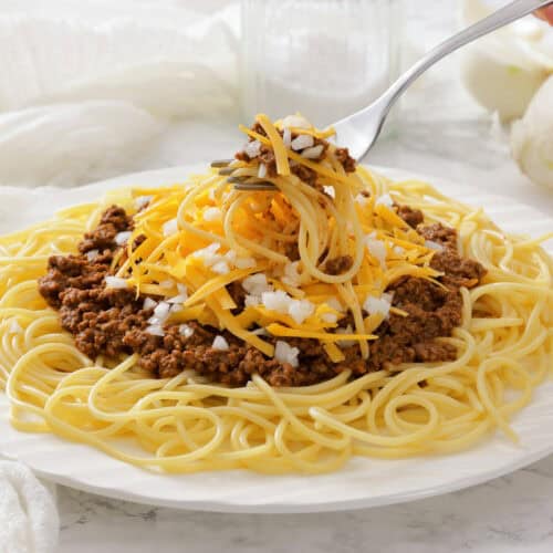 A plate of spaghetti topped with Cincinnati chili, shredded cheddar cheese, and chopped onions, with a fork lifting a bite.