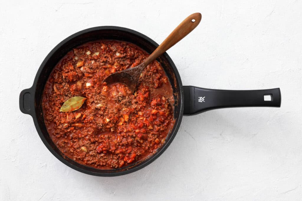 A black saucepan filled with tomato-based meat sauce, visible diced vegetables, a bay leaf, and a wooden spoon.