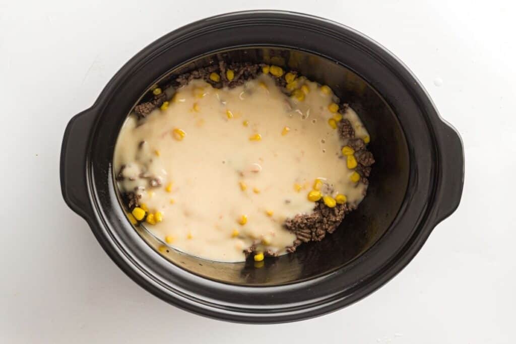A slow cooker filled with ground beef, corn, and a creamy soup mixture, viewed from above on a white background.