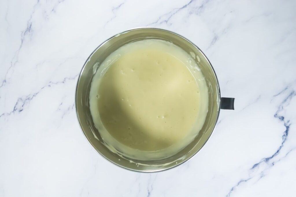 A metal mixing bowl filled with smooth, light yellow batter sits on a white marble surface.