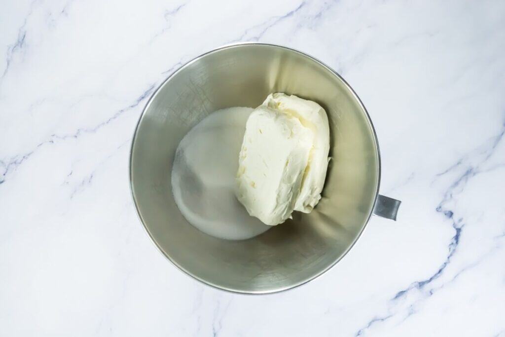 A metal mixing bowl containing cream cheese and granulated sugar on a white marble surface.