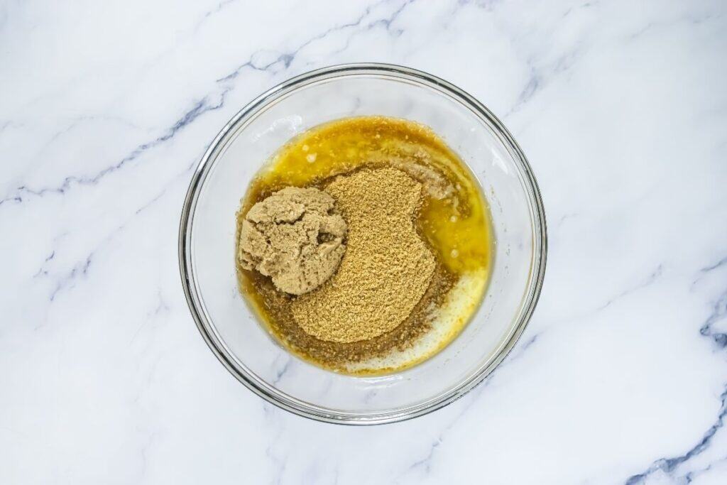 A glass bowl on a marble surface containing melted butter, brown sugar, and granulated sugar, ready to be mixed.