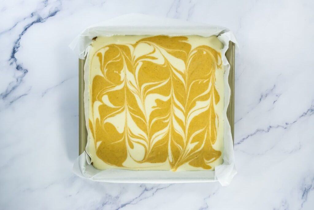 A square baking pan lined with parchment paper contains a swirled mixture of pumpkin and cream cheese batter on a marble surface.
