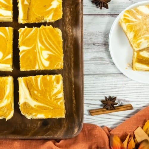 Six pumpkin swirl cheesecake bars are arranged in two rows on a wooden tray, displaying a marbled orange and white pattern on top.