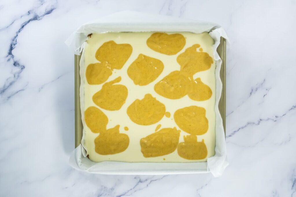 A square baking pan lined with parchment paper containing an unbaked layered mixture of white and yellow batter on a marble surface.