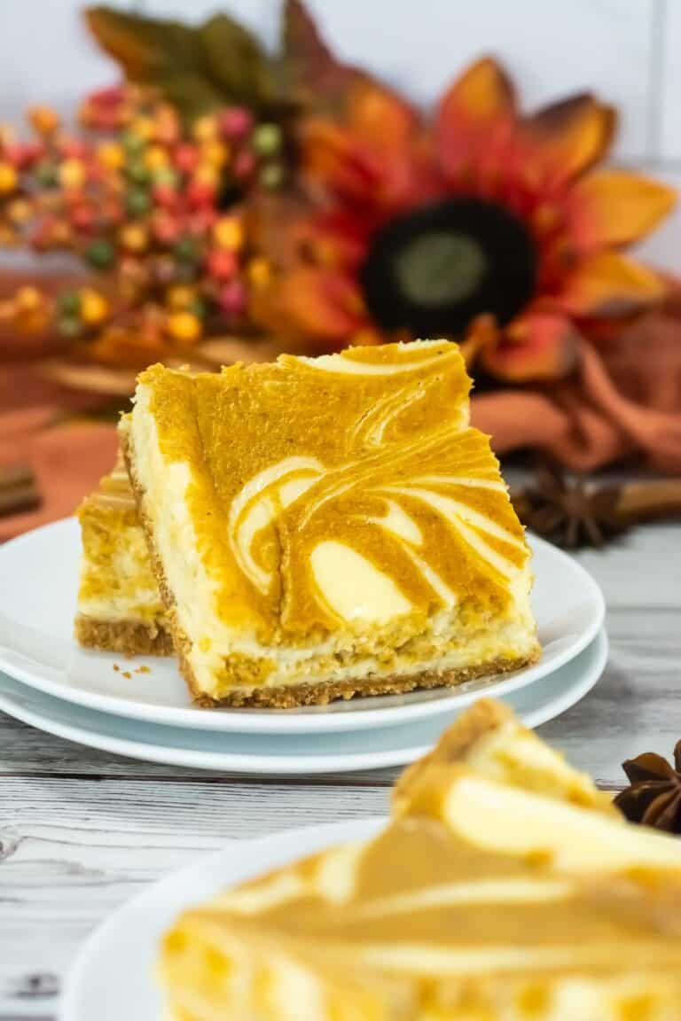 A slice of pumpkin cheesecake bar with a marbled swirl design on top, served on a white plate with autumn-themed decorations in the background.