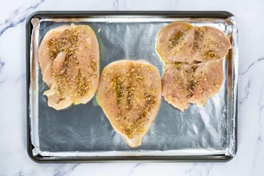 Raw chicken breasts seasoned with garlic and spices on baking sheet.