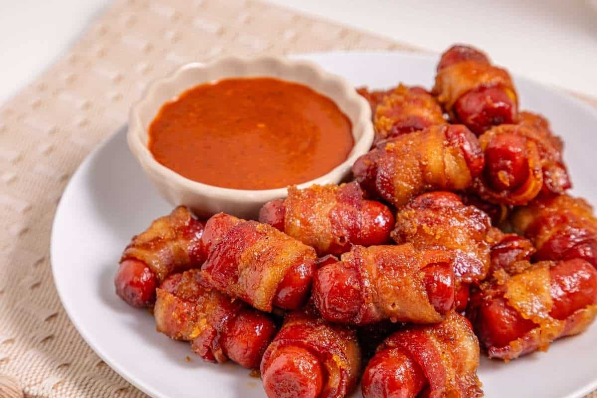 Plate of bacon-wrapped smokies caramelized and crispy, served with a dipping sauce on the side.