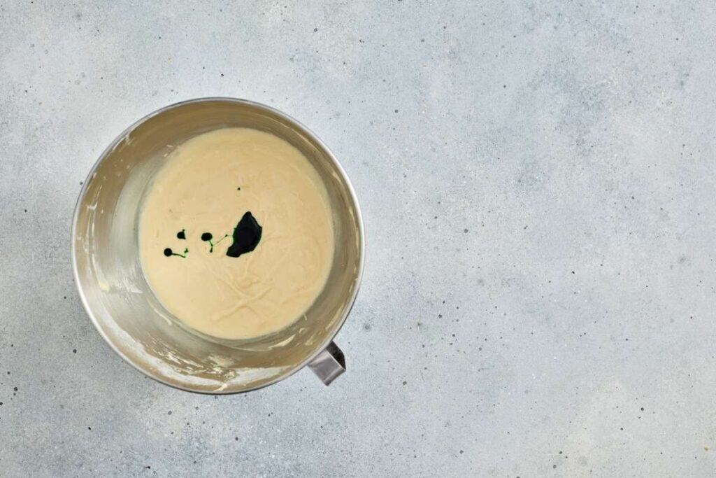 A stainless steel cup filled with mixture for cupcake with green gel food coloring.