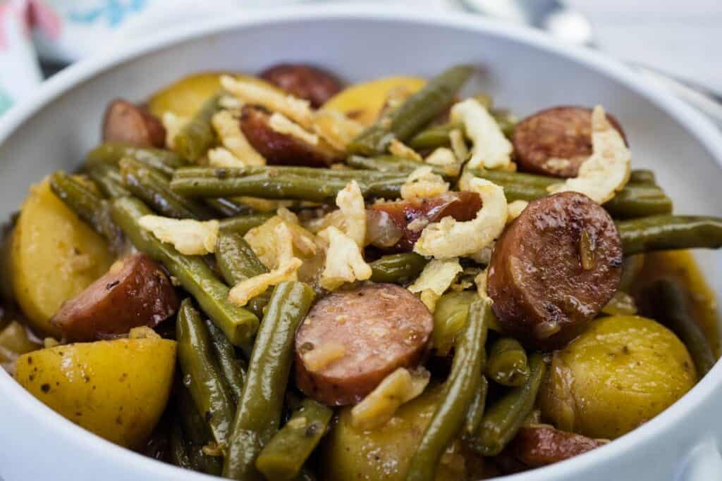 A bowl of Swamp Potatoes with cooked green beans, sliced sausage, and halved potatoes, topped with crispy fried onions.