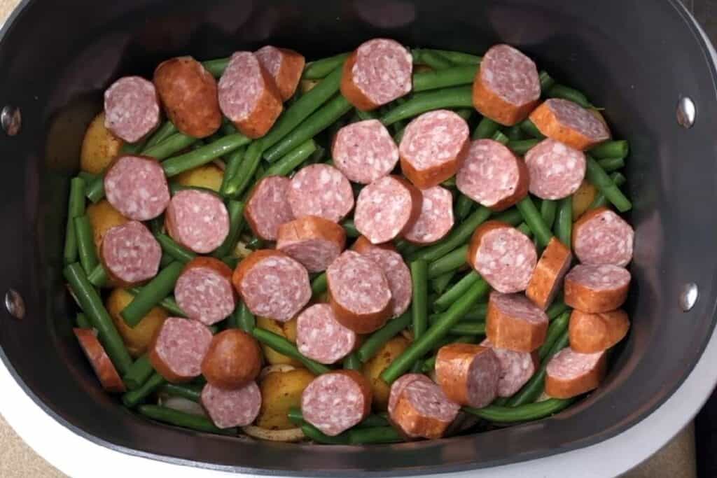 The image shows a slow cooker filled with sliced sausages, green beans, potatoes, and carrots, ready to be cooked into a flavorful stew.