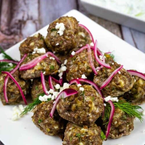 A white square plate of lamb meatballs toped with onion and fresh dill sits beside a small plate of tzatziki.