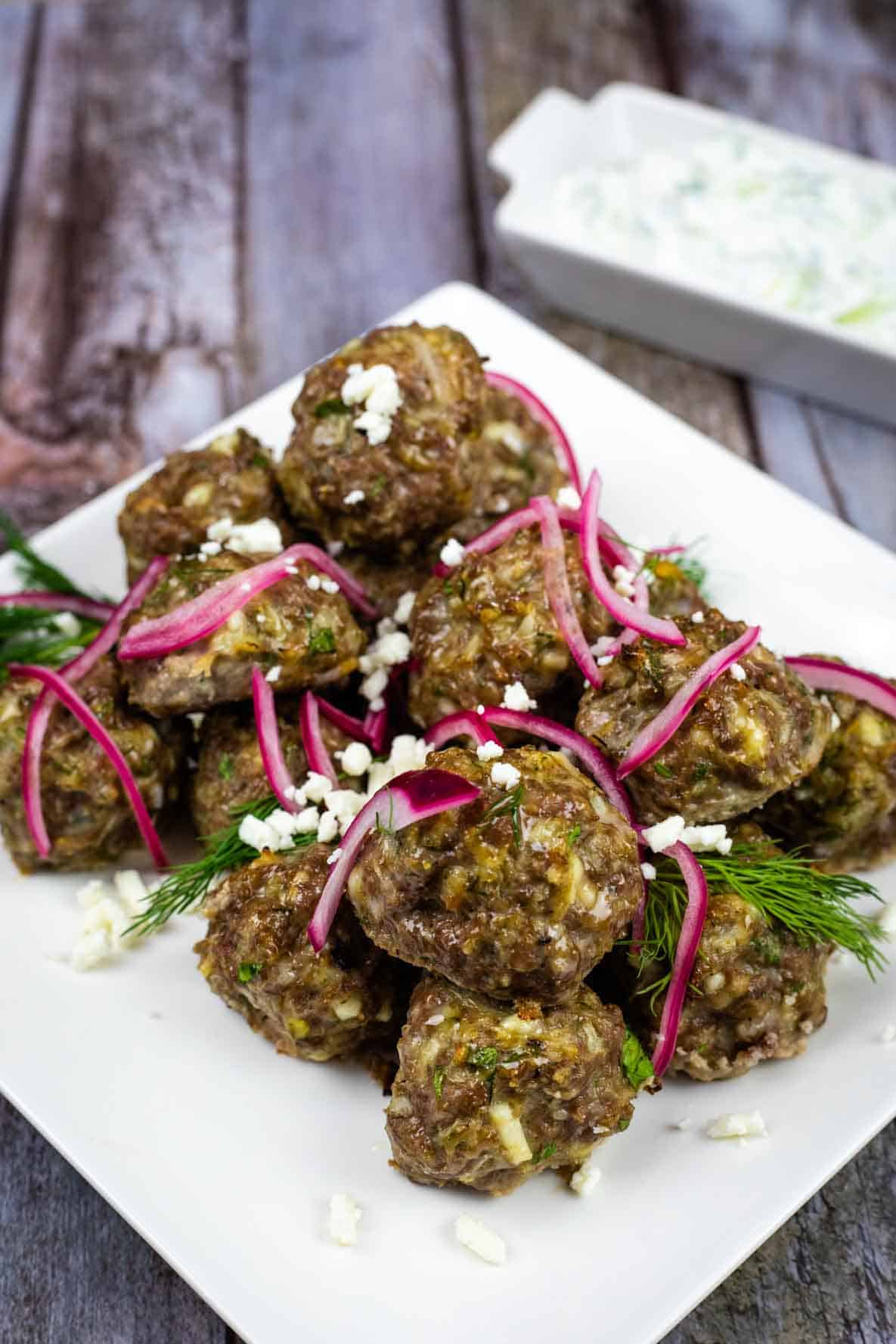 A white square plate of lamb meatballs toped with onion and fresh dill sits beside a small plate of tzatziki.