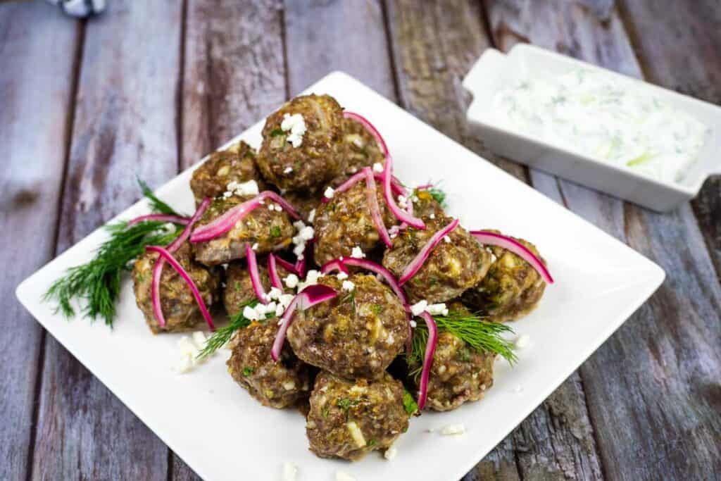 A white square plate featuring lamb meatballs sits beside a small plate of tzatziki.