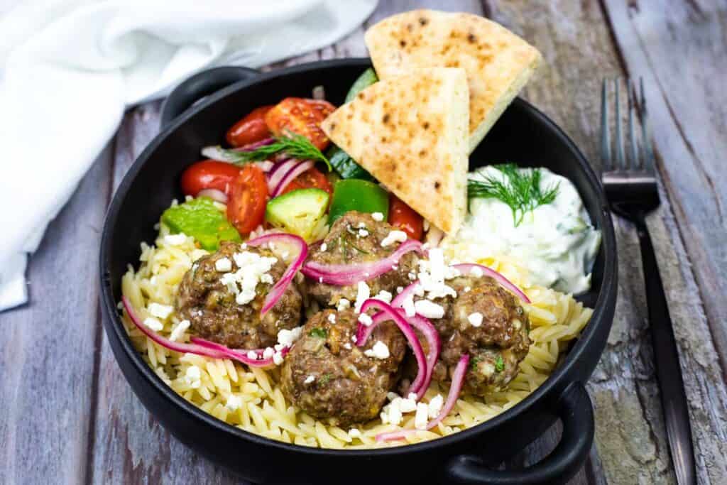 A bowl of lamb meatballs served with tzatziki, accompanied by rice and bread, rests beside a fork.