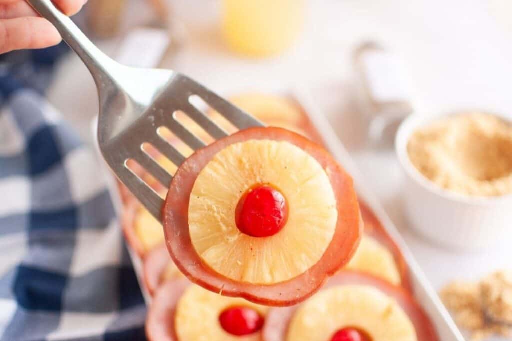 A spatula holding a slice of glazed ham taken from a plate.