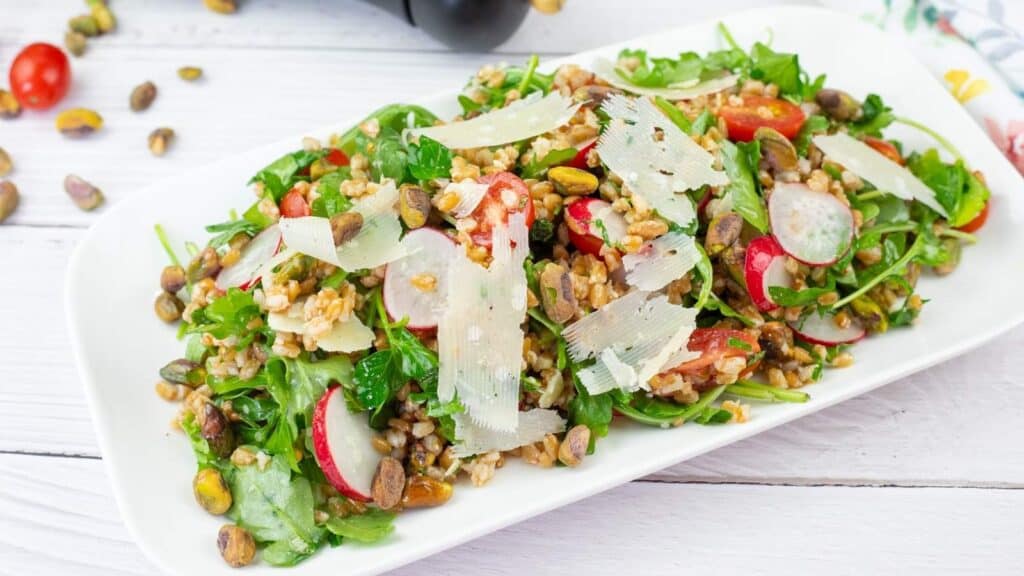 A white plate of Copycat Charlie Bird&rsquo;s Farro Salad topped with a flaky salt is placed on a wooden table.