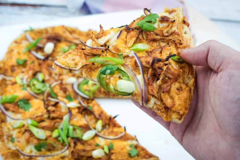 A hand holds a slice of pizza topped with shredded chicken, jalape&ntilde;o slices, red onions, and green onions; the rest of the pizza is visible on a white surface.