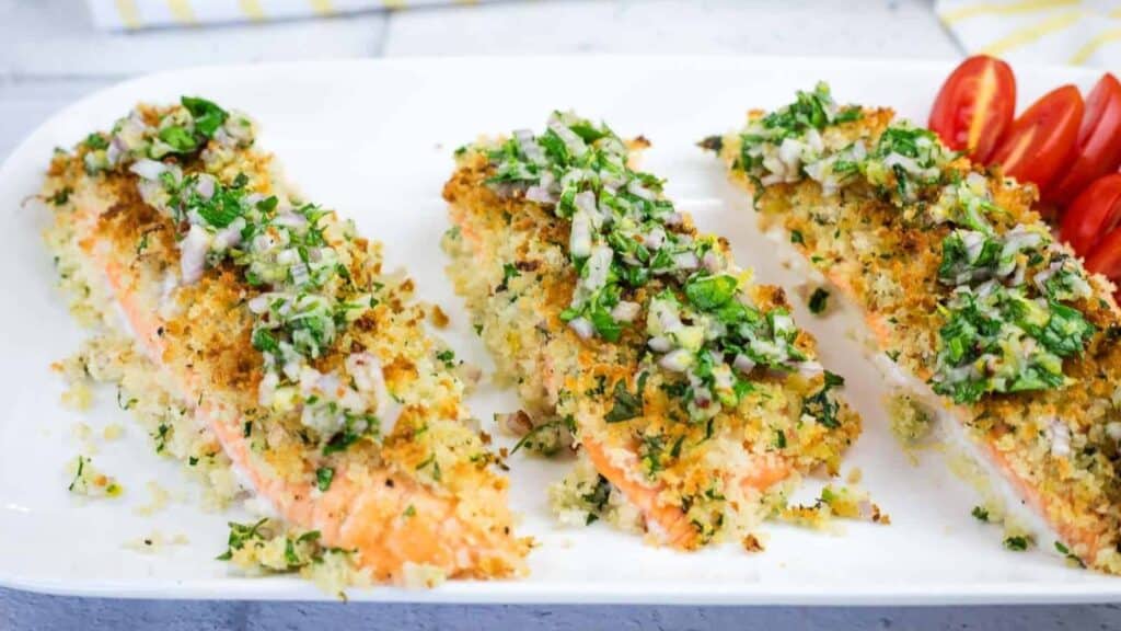 Three pieces of breadcrumb-crusted baked salmon topped with chopped herbs and served on a white plate with sliced cherry tomatoes.