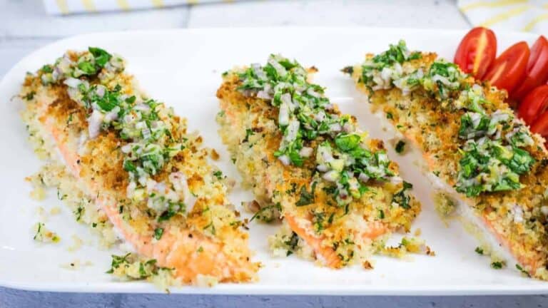 Three pieces of breadcrumb-crusted baked salmon topped with chopped herbs and served on a white plate with sliced cherry tomatoes.