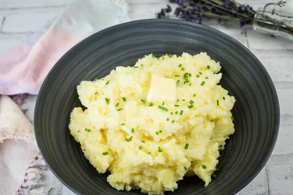 A black plate with mashed potatoes topped with butter and chopped chives.