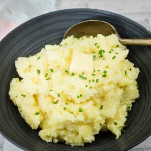 A black plate with mashed potatoes topped with butter and chopped chives beside a spoon.