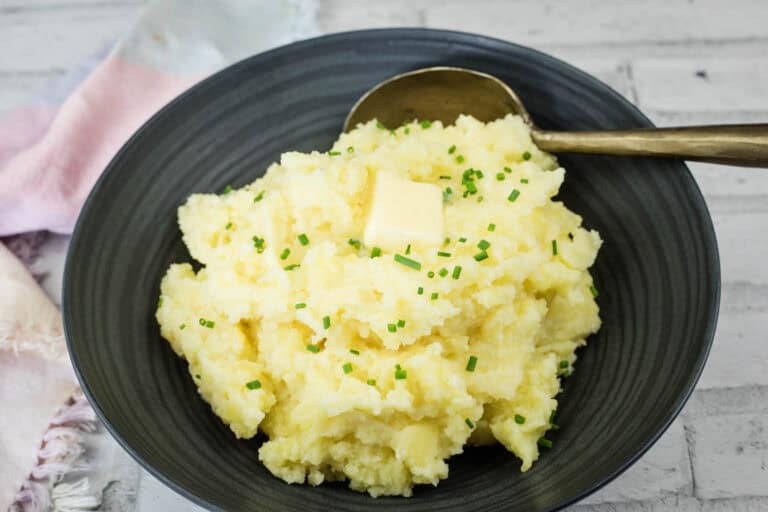 A black plate with mashed potatoes topped with butter and chopped chives beside a spoon.