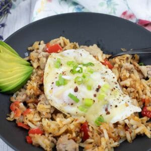 A black plate holds a Turkey Sausage Breakfast Bowl, accompanied by sliced avocado, topped with an egg and green onions.