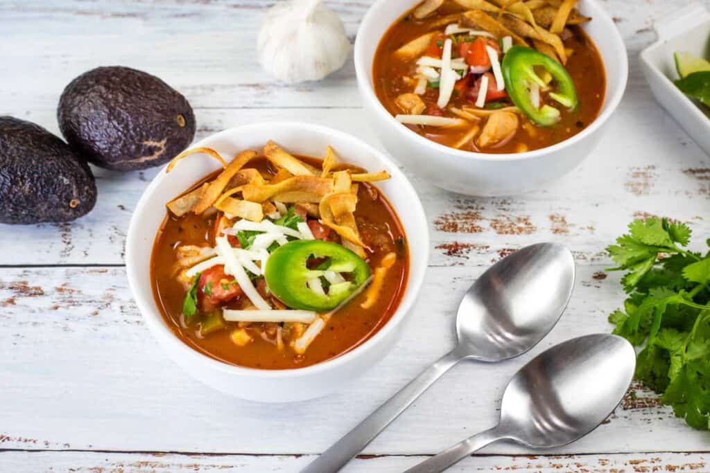 Two bowls of Chicken Tortilla Soup with two spoons, avocado, and garlic on the table.